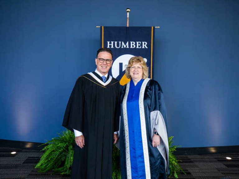 Humber president Ann Marie Vaughan posing with honorary degree recipient Anthony Longo in front of Humber flag