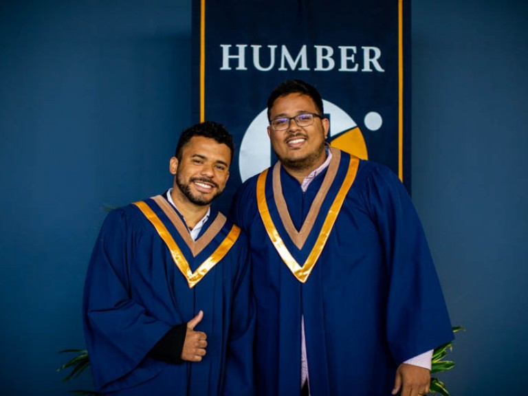 Two male graduates posing for photo in front of Humber flag, one holding thumbs up