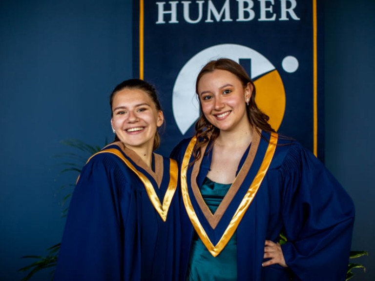 Two graduates pose for photo in front of Humber flag