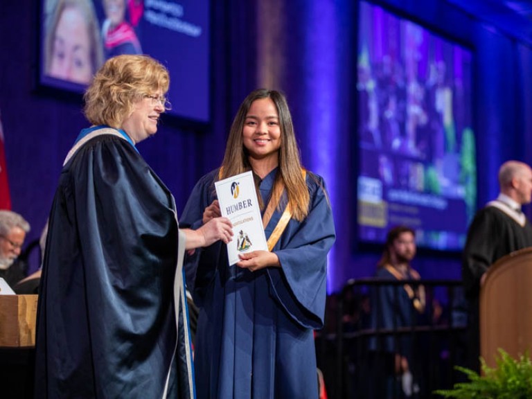 Graduate on stage holding certificate beside Ann Marie Vaughan