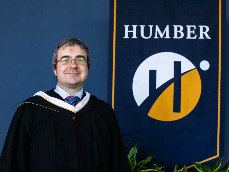 Honorary degree recipient Andrew Monkhouse poses for photo beside Humber flag 