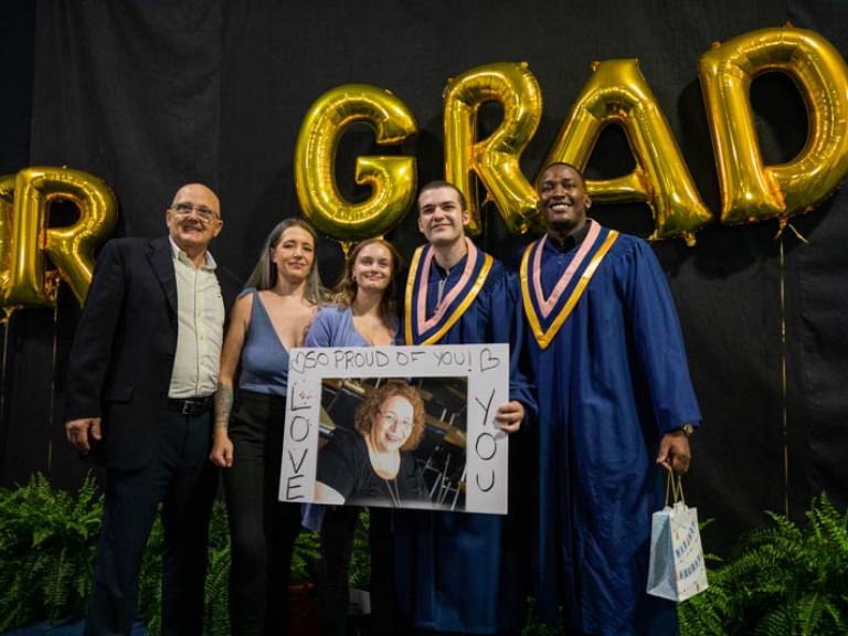 Two graduates taking photo with three guests in front of gold grad balloons