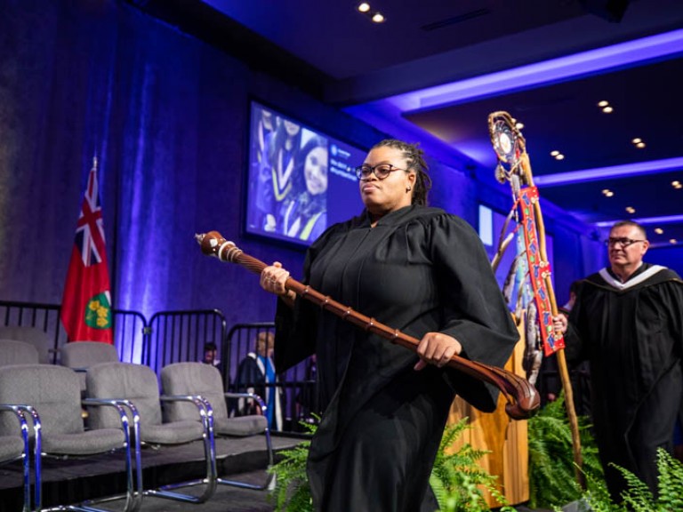 Two people carrying Indigenous staffs walk on stage