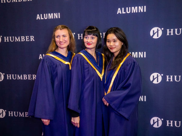Three graduates take photo in front of Humber alumni wall