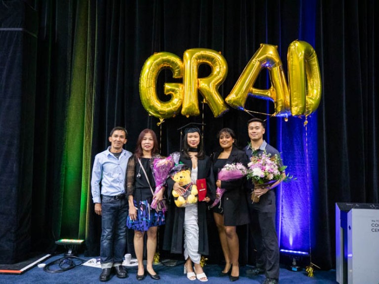 Graduate takes photo with four guests underneath gold GRAD balloons