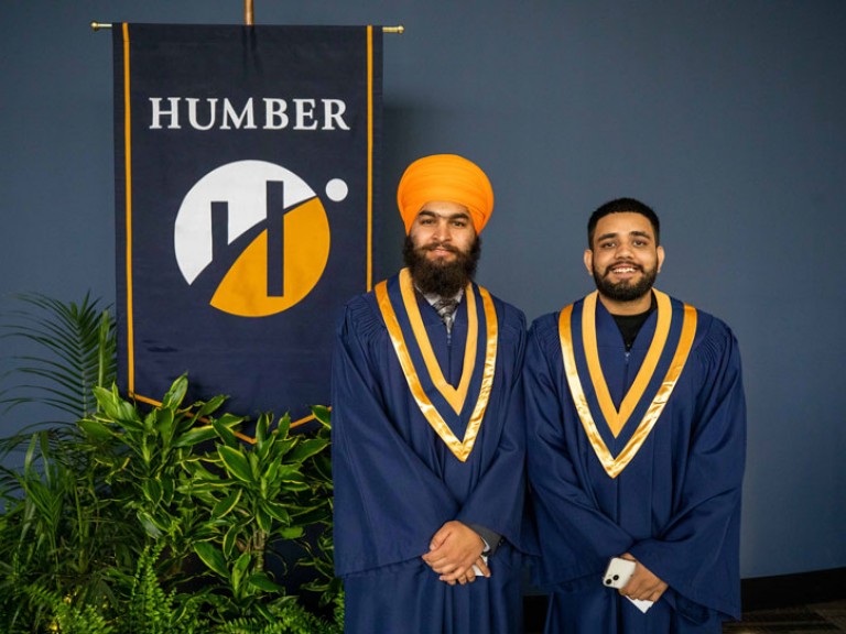 Two graduates pose for photo in front of Humber flag