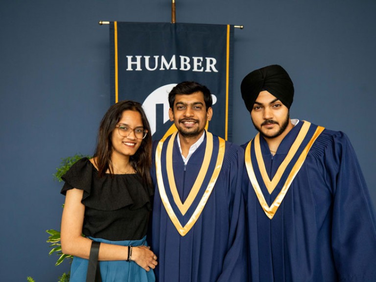 Two graduates and guest pose for photo in front of Humber flag