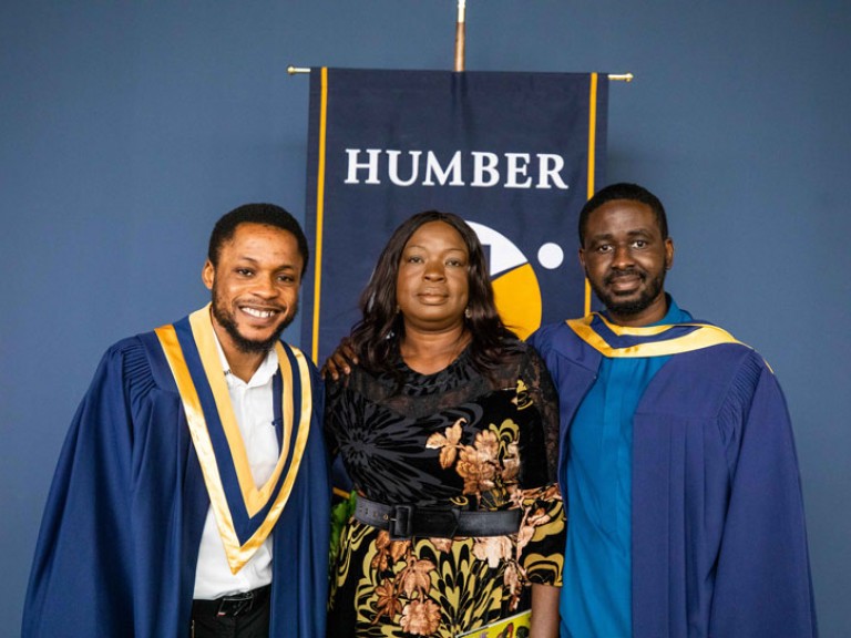 Two graduates take photo with guest in front of Humber flag