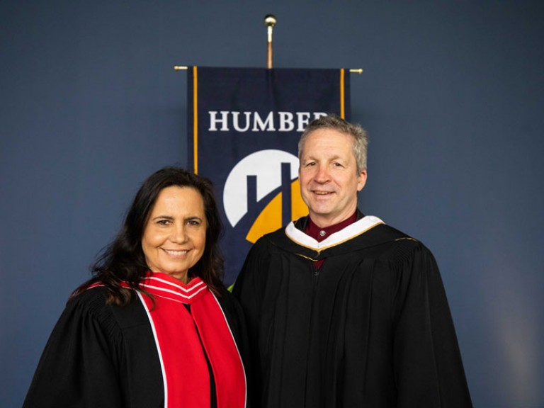 Honorary degree recipient Jim Estill poses with faculty member in front of Humber flag