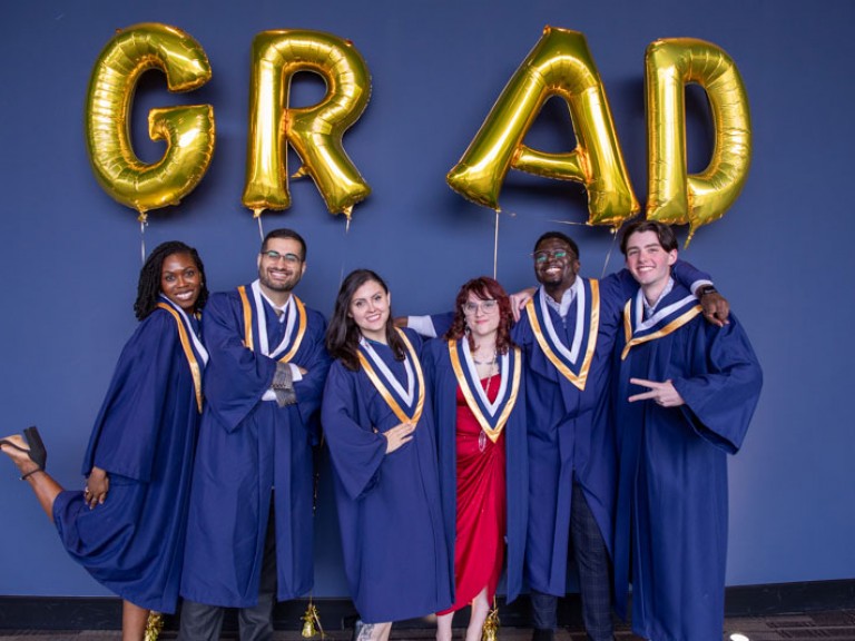 Six graduates posing for photo under gold GRAD balloons