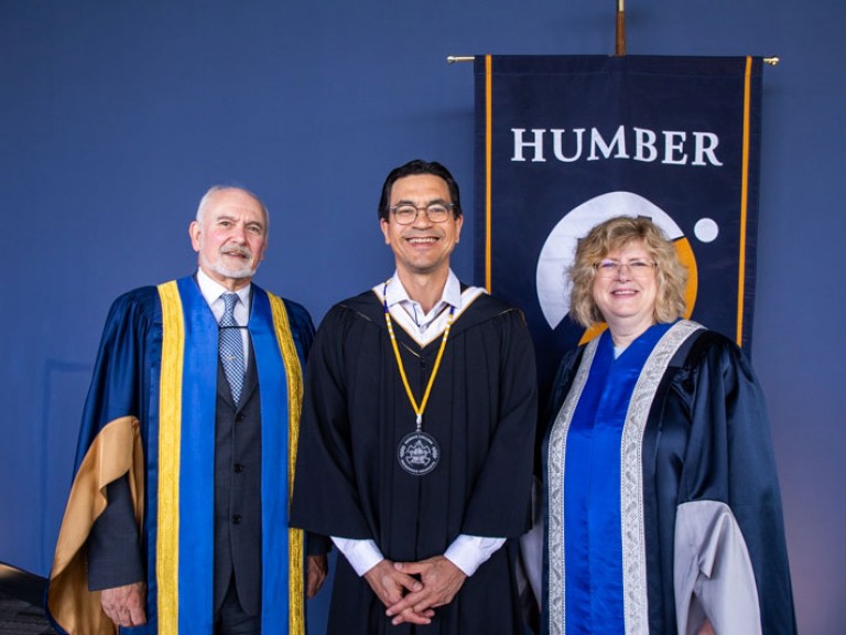 Humber president and honorary degree recipient pose with another Humber faculty member in front of Humber flag