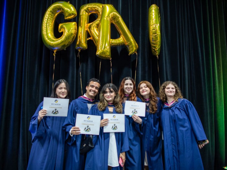 Six graduate pose under GRAD balloons
