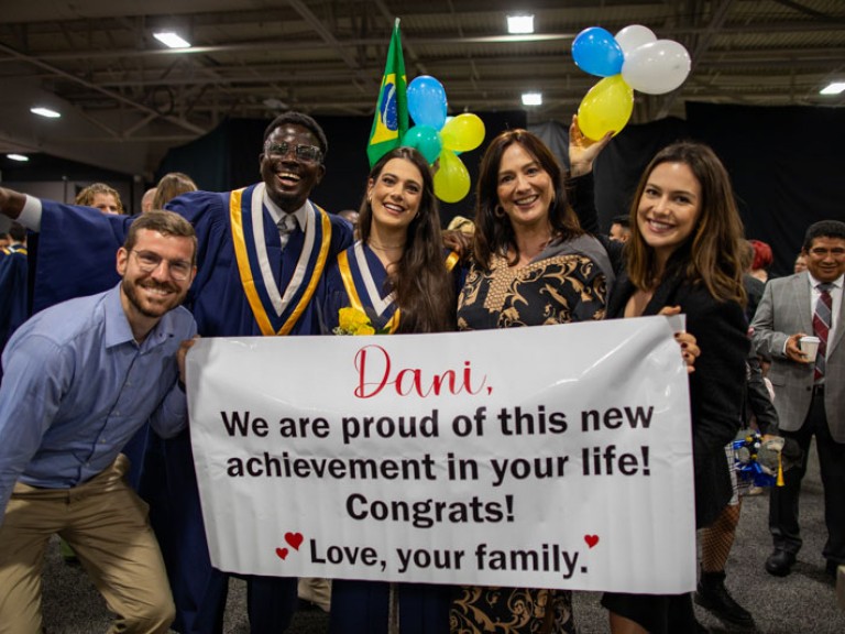 Two graduates and three ceremony guests hold sign that reads Dani, We are proud of this new achievement in your life! Congrats, Love, your family.