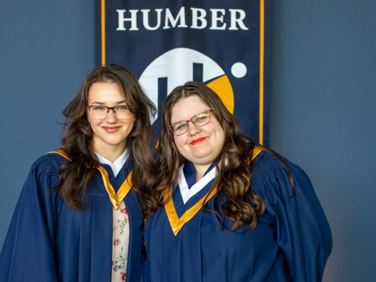 Two graduates take photo together in front of Humber flag