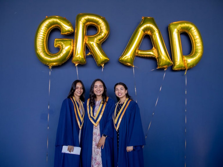 Three graduates smile for photo beneath GRAD balloons