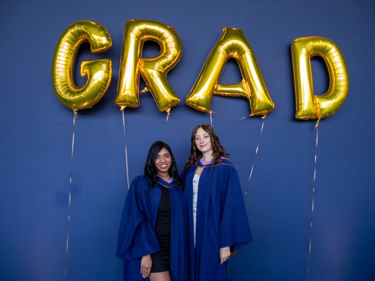 Two graduates pose for photo beneath GRAD balloons