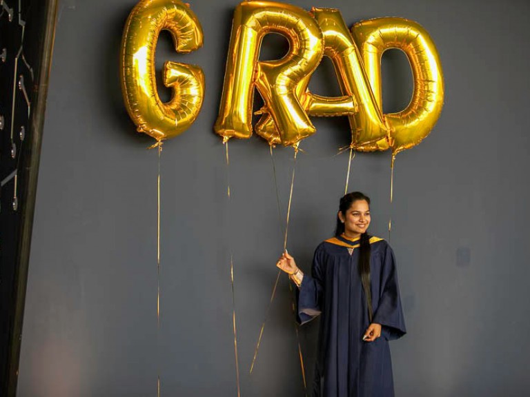 Graduate takes photo underneath gold GRAD balloons