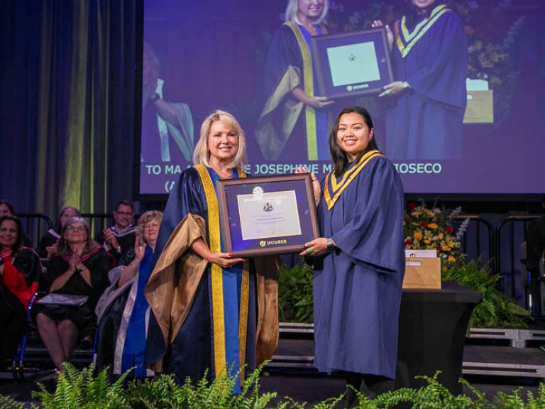 Graduate holds up framed award with Humber faculty member