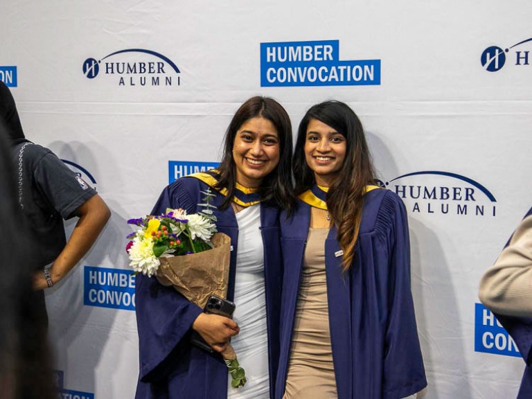 Two graduates take photo together in front of Humber convocation wall