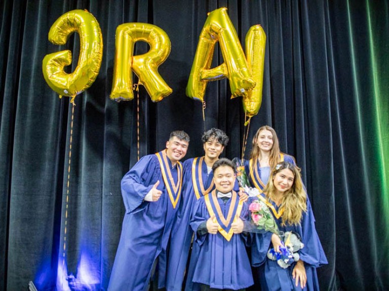 Five graduates take photo together under gold GRAD balloons