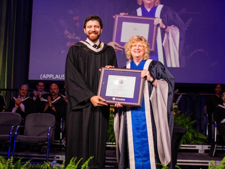 Honorary degree recipient Tareq Hadhad holds framed award with Humber president on stage