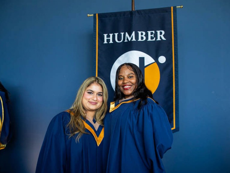 Two graduates smile for camera in front of Humber flag
