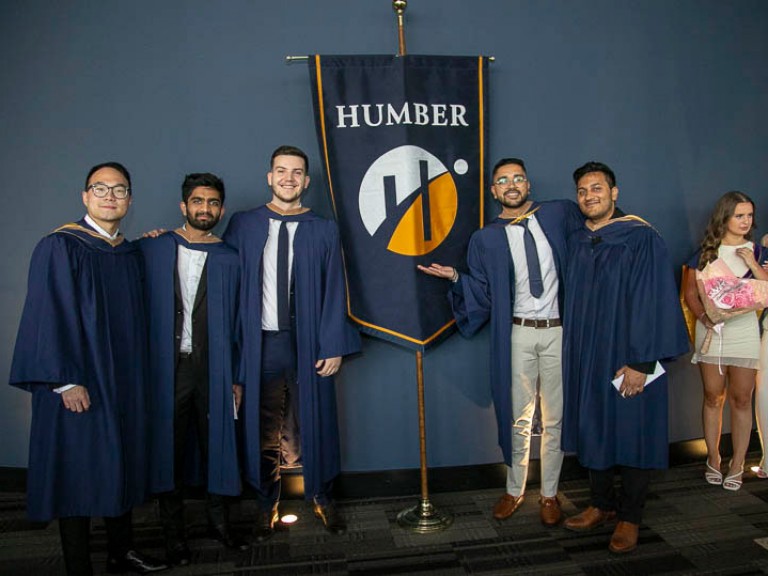 Five graduates pose beside Humber flag