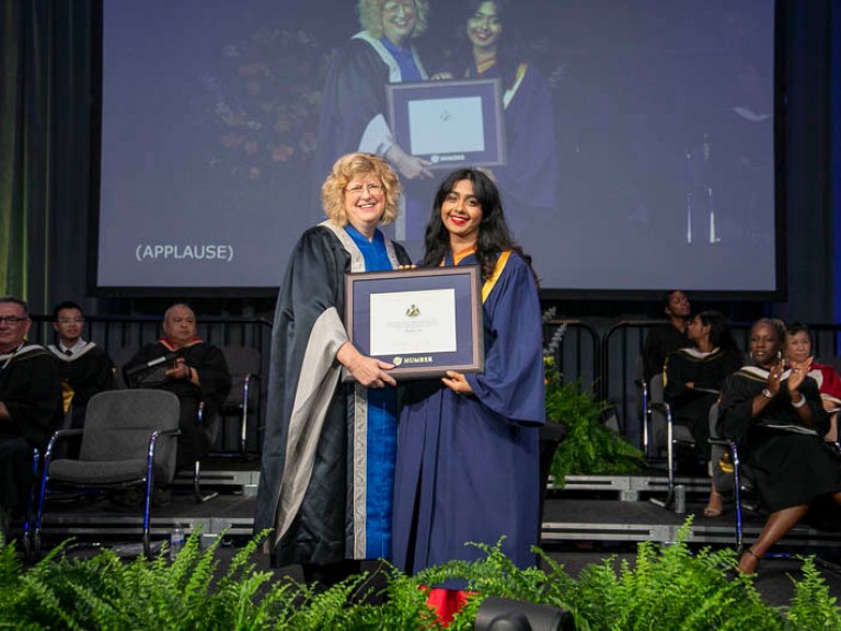Graduate holds framed award with Humber president on stage
