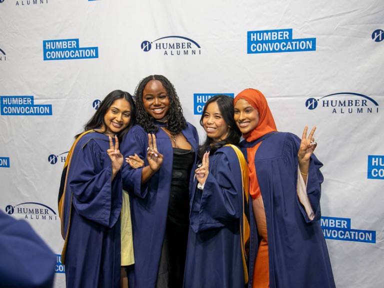 Four graduates hold peace signs as they pose in front of Humber Convocation wall