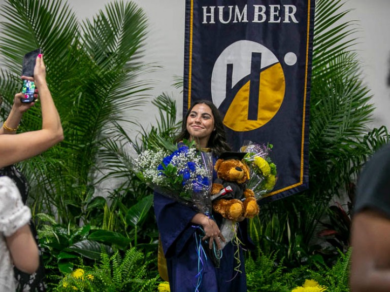 Graduate holds flowers and teddy bear as someone takes a cell phone picture