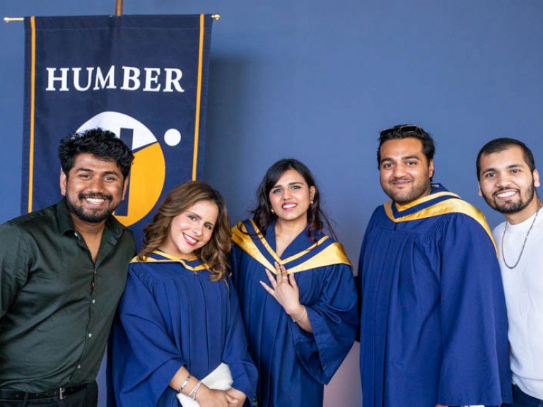 Three graduates and two guests take photo in front of Humber flag