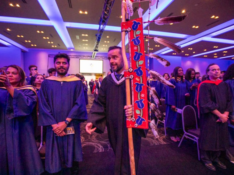 Faculty member proceeds into hall holding ceremonial Indigenous staff