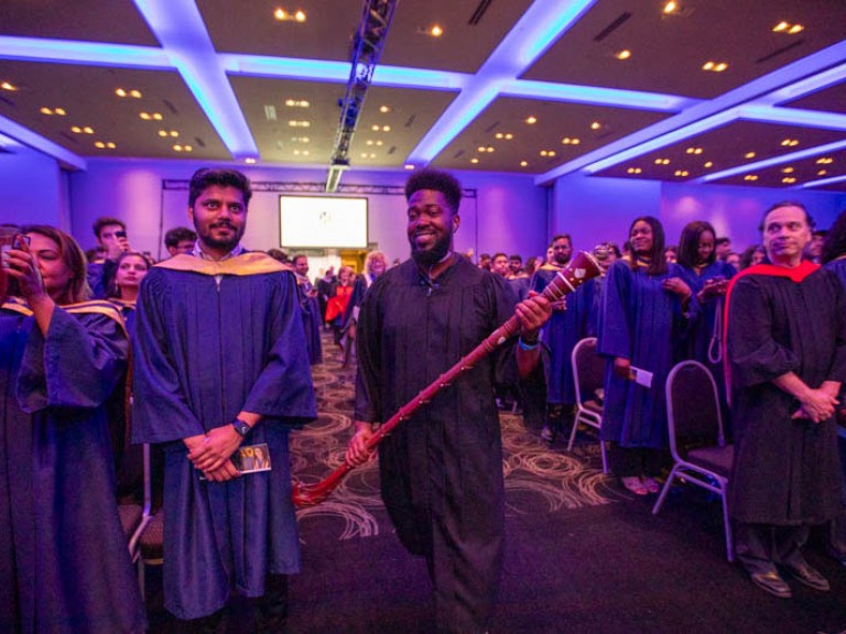 Faculty member proceeds into hall holding ceremonial Indigenous staff