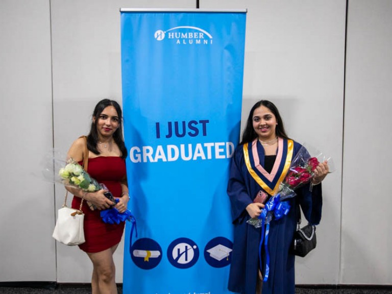 Two graduates holding flowers stand beside banner that reads I Just Graduated