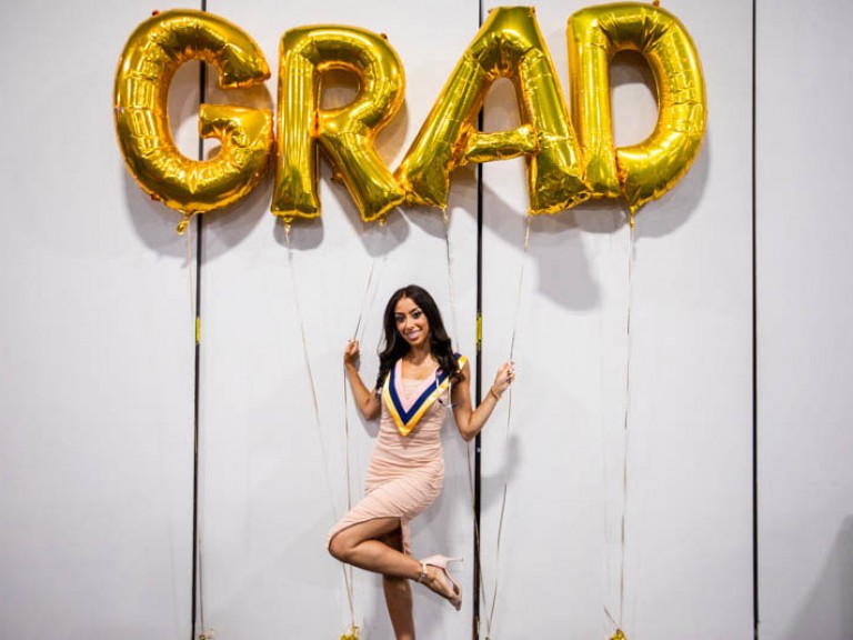 Graduate poses underneath gold GRAD balloons