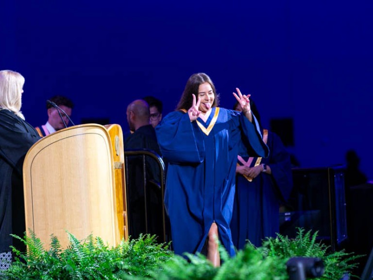 Graduate holds up two peace signs with their fingers as the cross stage