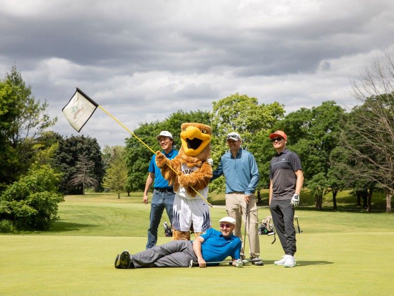 Four people pose on course with mascot, one person lying on the ground in front
