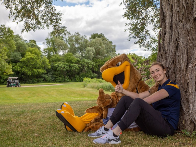 Person with Humber mascot sitting at base of tree holding thumbs up