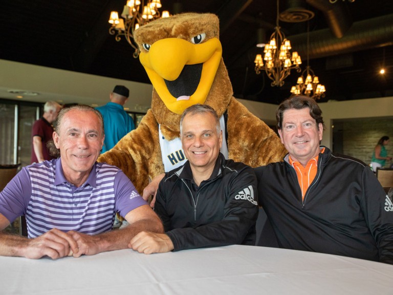 Three people sitting at table with Humber mascot behind them
