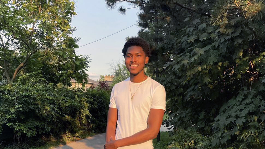Jorien Stephens smiles in the sun, standing on a paved road surrounded by trees. He looks relaxed and happy.