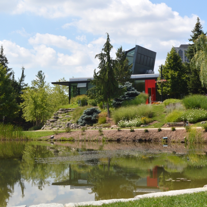A glass building with a red door sits by a large pond surrounded by trees