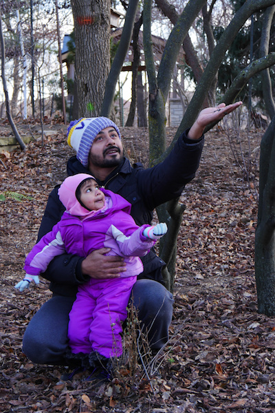 A toddler in a snowsuit watches as her father holds up a hand filled with bird seeds.