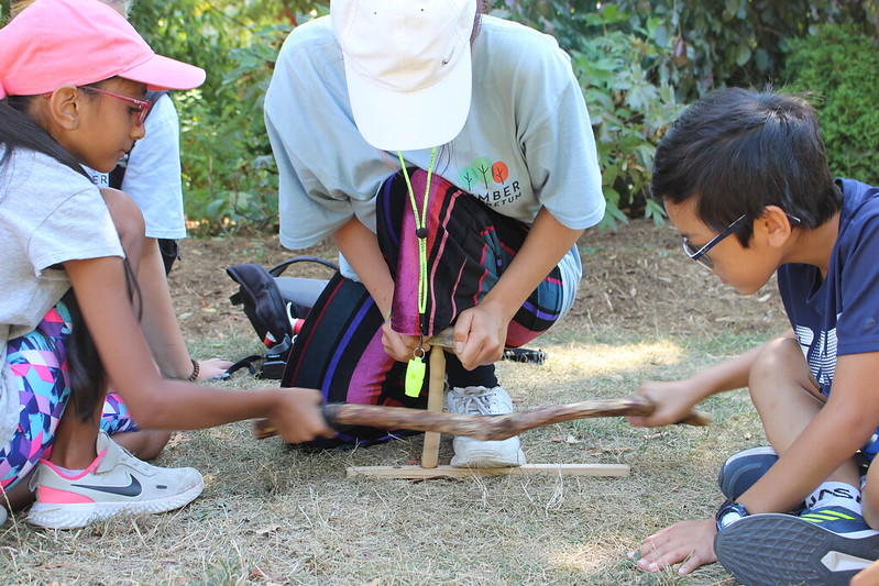 A counsellor helps two campers trying to start a friction fire by rubbing sticks together