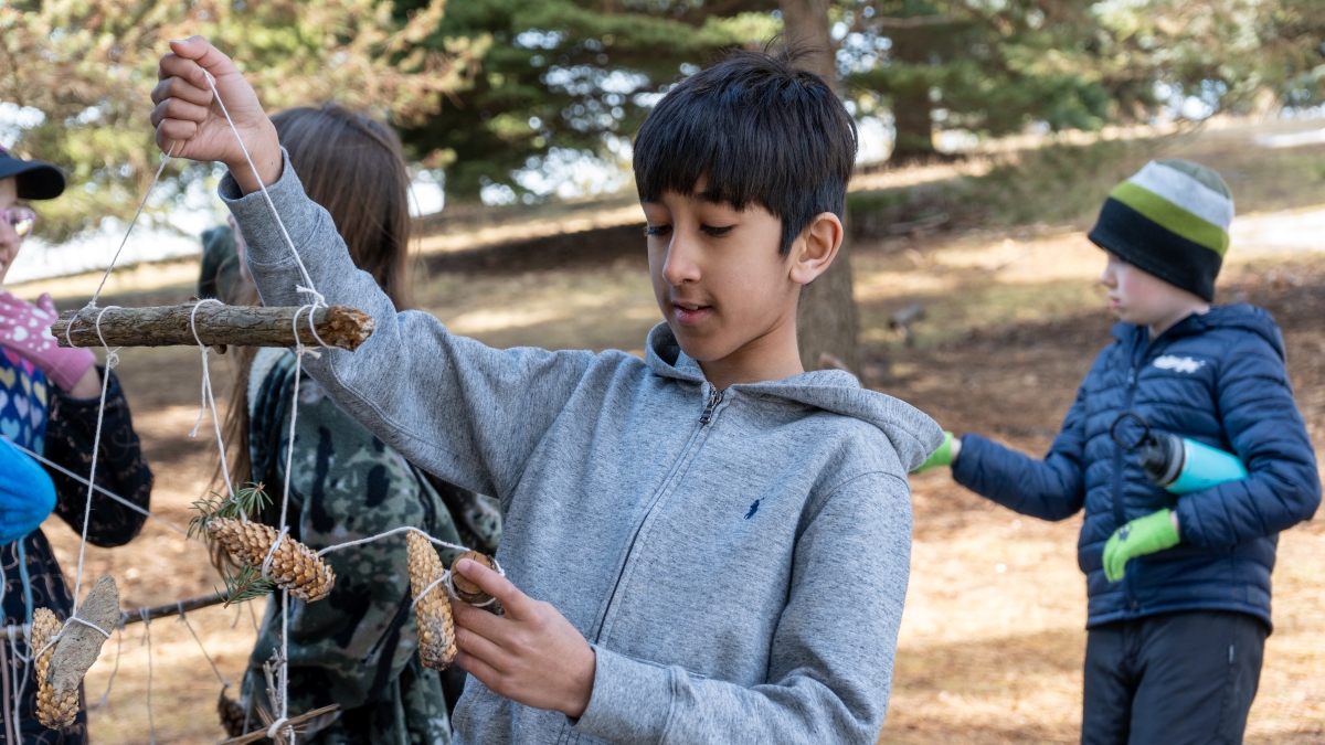 Standing outside a child looks at a mobile they've made out of natural materials