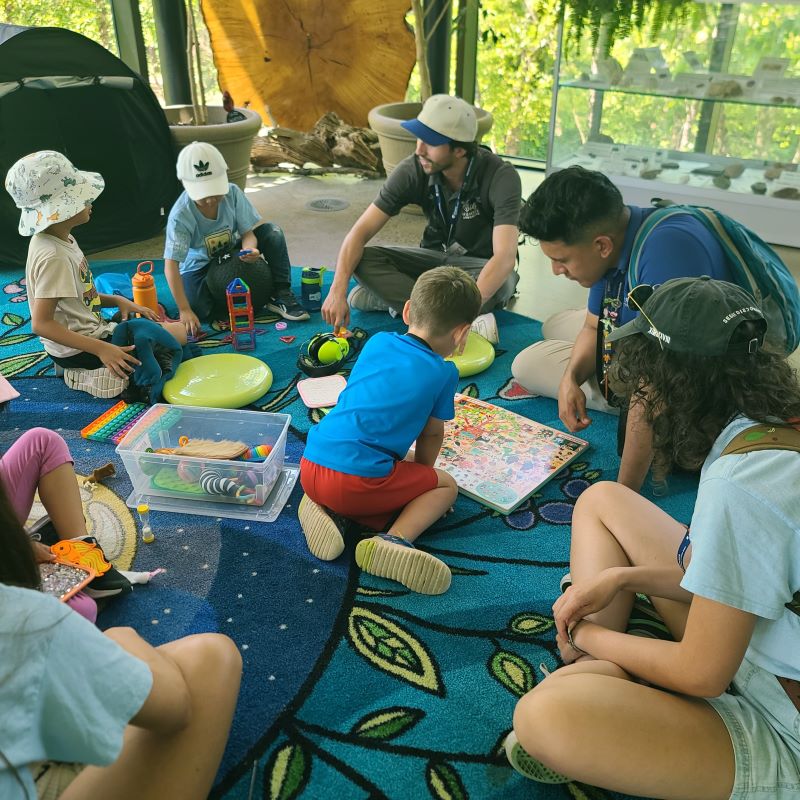 A group of children sit indoors on a carpet with camp leaders, playing with puzzles and toys.  