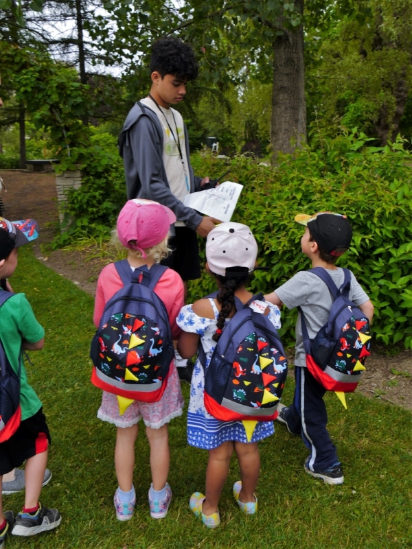 Outside in the garden of the Humber Arboretum, a teen gives instructions to a group of young campers in matching backpacks
