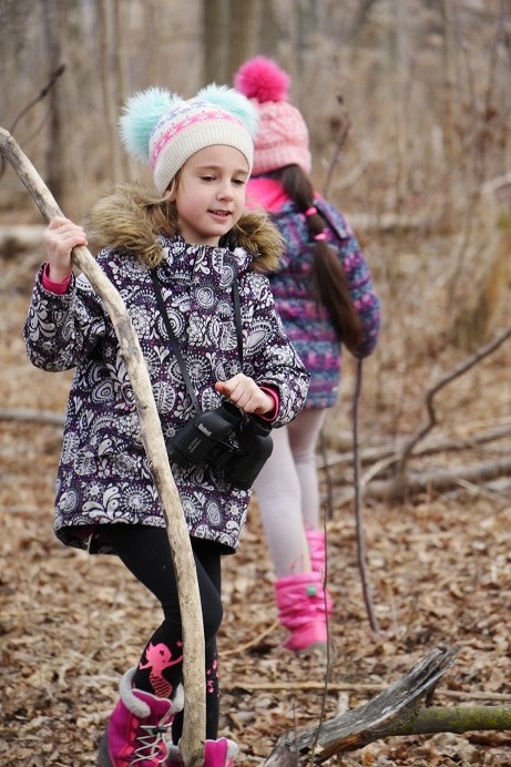 A girl in winter clothes walks through a wooded area using a long branch as a walking stick.