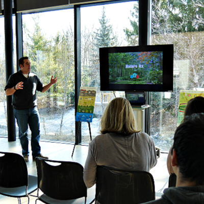 A man gives a presentation in front of floor-to-ceiling windows that provide a view of trees outside