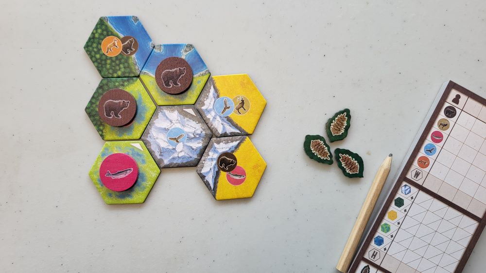 Hexagon tiles with different habitats are laid out on a table next to a score sheet. Some of the tiles have animals tokens on them such as a bear.