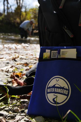 A Water Rangers branded test kit pouch leans against a backpack at the edge of the Humber River. In the background, an individual in an Arboretum staff t-shirt stands bent over in the river.